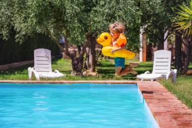 Yüzme şortu giymiş heyecanlı bir çocuk, sarı ördek şamandırası takıyor, yaz tatillerinde sakin güneşli bir havuza atlıyor.