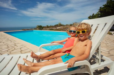 Bir erkek ve bir kız, yaz tatillerinde mavi gökyüzünün altında, güneşli bir havuz kenarında, renkli güneş gözlüğü takarak güneşleniyorlar.
