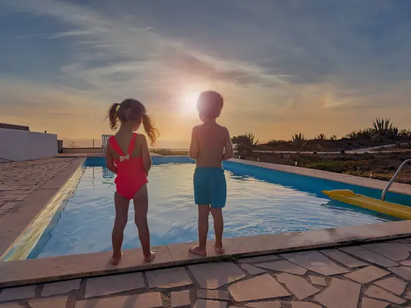 2人の子供がプールの端に立って 夕日を眺めながら後退する太陽の光によって概説された数字を見ます — ストック写真