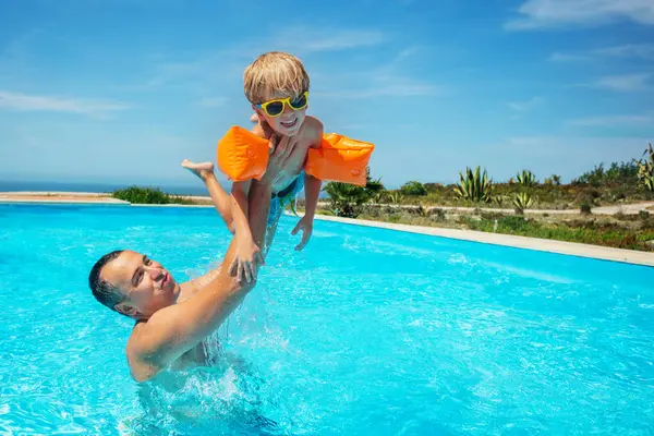 遊び心のある方法で水の上に子供を持ち上げるスイミングプールでは 泳ぐのを助けるために腕に浮かぶサングラスを身に着けている少年 — ストック写真