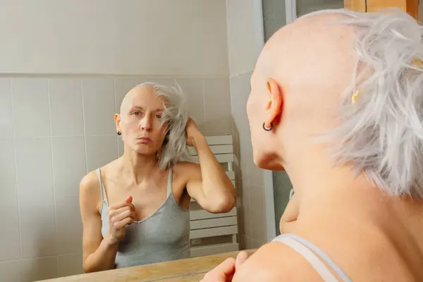 彼女の外観を考えると 清潔な頭を持つ悲しい若い女性は鏡に直面しながら灰色のウィッグを保持します — ストック写真