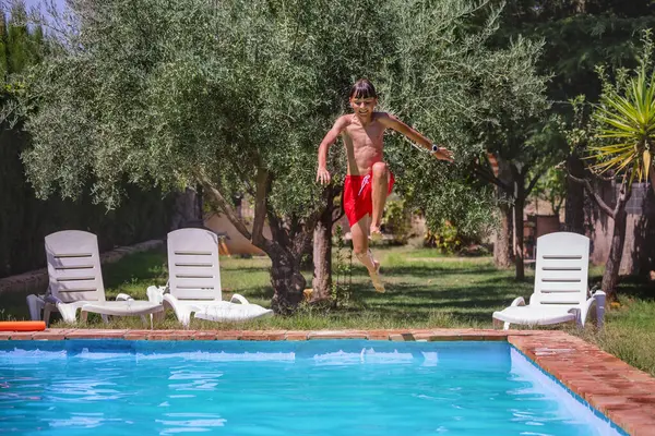 Captured in mid-air, a child sporting a pair of red swim shorts is diving into an aqua pool, flanked by white deck chairs and shaded by verdant trees.