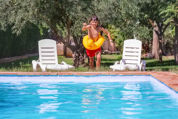 A young boy in red swim shorts is joyfully preparing to jump into a swimming pool, holding a yellow inflatable ring