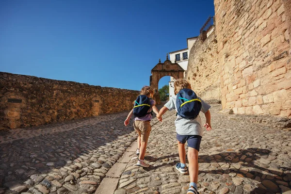 Kids tourists run holding hands in old part of Ronda, enjoy their summer vacations in the South of Spain, view from behind