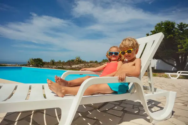 Cute boy and girl, two siblings lounging on white sunbeds enjoy the summer sun, relaxing on pool chairs with resort\'s swimming area on background