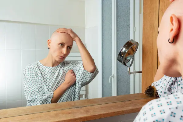 深く考え 髪のない患者は トイレの鏡で自分自身を見つめ 病院のドレスを着ています ストック写真