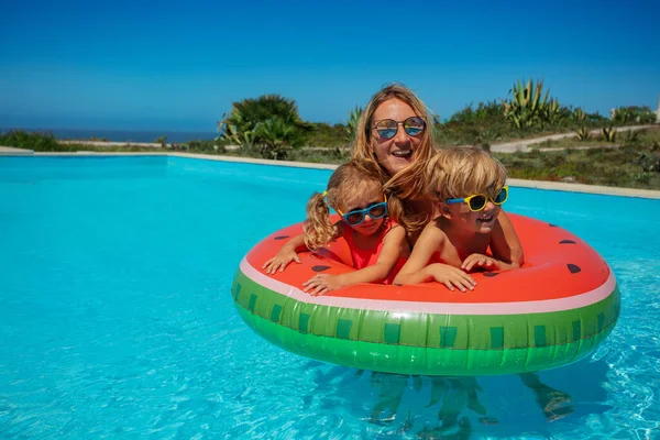 戴着太阳镜 笑着和抱着两个年幼孩子的女人在充气西瓜里游泳 图库照片