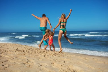 Genç bir aile kumlu sahilde neşeli bir atlayışın keyfini çıkarıyor. Arka planda dalgalar çarpıyor, mutlu tatiller.