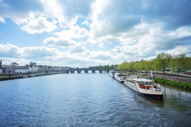 Hollanda, Maastricht 'te bulutlu bir gökyüzüne karşı kayıklar ve taş bir Servaas köprüsü ile Meuse nehrinin manzarası.