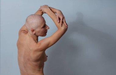 Çıplak gövdeli kel bir genç kız kollarını açık gri bir arka plana doğru uzatıyor.