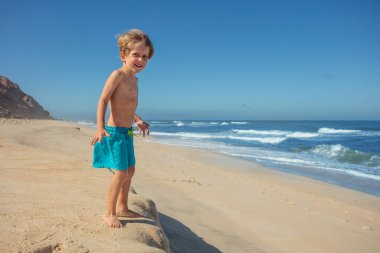 Kumlu sahilde, küçük bir çocuk çıplak ayakla denizi gözlemler, arkasında kayalık bir kayalık ve engin mavi bir gökyüzü ile yüzmeye hazırdır..