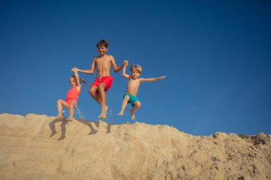 Üç neşeli çocuk açık mavi gökyüzüne doğru kum tepeciklerinden atlıyor