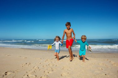 Çocuklar kıyı şeridi boyunca neşe içinde koşarlar, parlak güneşin altında kumların üzerinde ayak izleri bırakırlar, yaz tatillerinde eğlenirler.