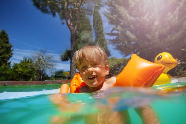 Turuncu kollu neşeli bir çocuk güneşli bir günde turkuaz bir yüzme havuzunda oynuyor ve arka planda şişme bir ördek var.