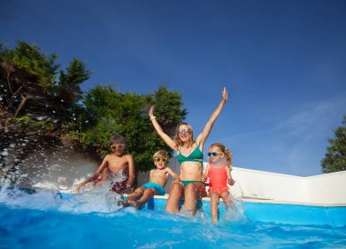Çocuklar ve gülümseyen bir kadın renkli yüzme kıyafetleri içinde el kaldırıyor, güneş gözlüğü takıyor ve havuz kenarında ağaçlarla vakit geçiriyorlar.