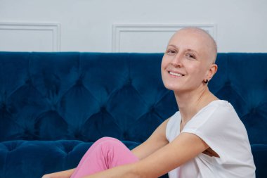 Kemoterapi tedavisi gören neşeli bir kadının gücü, beyaz bluzu ve pembe pantolonu olan, aydınlık bir odada, lüks, mavi bir kanepeye karşı gelişigüzel oturur.