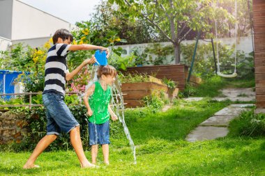 Gençler, güneşli bir bahçede yeşil gömlekli küçük bir çocuğa mavi kovadan neşeyle su dökerler.