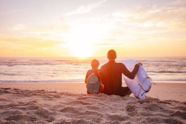 İki sörfçü, baba ve oğul sörf tahtasıyla kumun üzerinde oturur ve günbatımının okyanusun üzerine düşüşünü izlerler.
