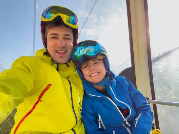 stock image Cheerful child with his father wearing ski gear enjoy a ride on a cable car do selfies against the sunny picturesque landscape of snowy mountains