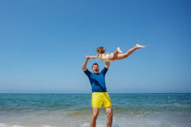 Bir plaj gününün tadını çıkarırken, bir baba ve kızı onu suyun yanında döndürürken gülüyorlar, baba heyecandan çığlık atıyor.