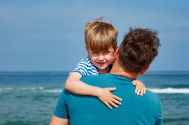 Kumsalda güneşli bir günde, babası onu tutarken mutlu bir çocuk sırıtır ve arka planda okyanus görünür.