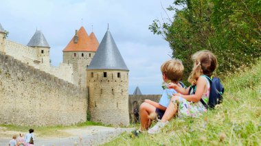 Sırt çantalı gezginler, Carcassonne 'un ortaçağ taş kulelerine bakan çimlerin üzerinde otururlar. Açık bir günde, Fransa seyahatlerinin tadını çıkarırlar.