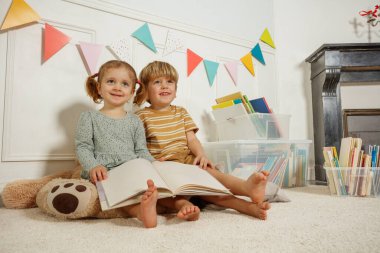 İki genç huzur içinde oturuyorlar, her biri kendi kitabına odaklanmış. Sahne, beyaz panelli bir duvara asılı canlı bir afişle tamamlanıyor.