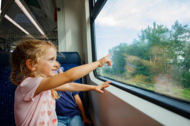 Küçük bir kız. Kıvırcık sarı saçlar ve kalp desenleri olan pembe bir gömlek hareket halindeki bir trenin penceresinden dışarısını işaret ediyor.