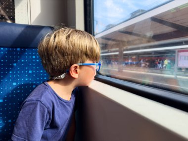 Mavi gözlüklü genç bir gezgin, yaz tatili boyunca dışarıdaki yoğun platformu gözetleyerek trenin camından dışarı bakıyor.