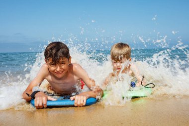 Two boys joyfully ride bodyboards as ocean waves crash over them on a sunny beach, kids all covered in splashes clipart