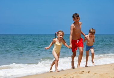 Çocuklar, kızlar ve erkekler kumlu sahilde koşuşturuyorlar, el ele tutuşuyorlar, arkada deniz var.