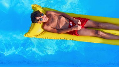 Safir havuzunda zahmetsizce süzülen genç bir insan, kırmızı yüzme elbiseleri ve koruyucu güneş gözlükleriyle güneşli bir kehribar şişme yatakta dinlenirken resmediliyor.