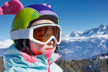 Karla kaplı dağların arkasında canlı kayak elbiseli, gözlüklü ve kanatlı komik kasklı bir kız var.