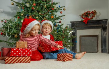 Kırmızı ve beyaz Noel Baba şapkaları takmış bir çift çocuk Noel ağacının yanında bir sevgi anı paylaşıyor.