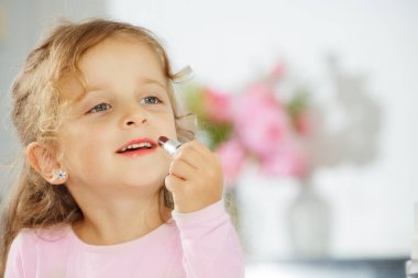 Sarı saçlı, pembe ruj sürmüş ve hafifçe aynaya bakıp gülümseyen küçük bir kız.