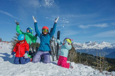 Neşeli bir grup çocuk ve renkli kayak ceketleri, miğferler giyen bir anne, karları havaya fırlatıyor, dingin mavi gökyüzüne karşı kurulmuş, nefes kesen bir dağ zeminine