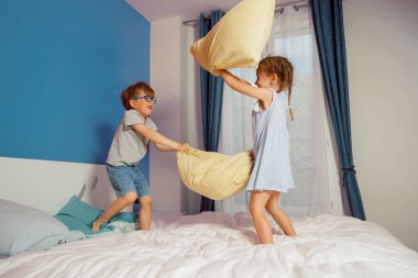 Two children engage in a playful pillow fight on a bed with blue walls and curtains in the background clipart