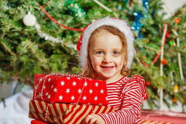 Noel Baba şapkalı neşeli bir çocuk Noel hediyelerini süslenmiş bir Noel ağacının önünde tutuyor.