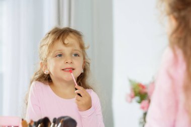 Genç bir kız aynadaki yansımasına bakarken dudak parlatıcısı sürüyor.