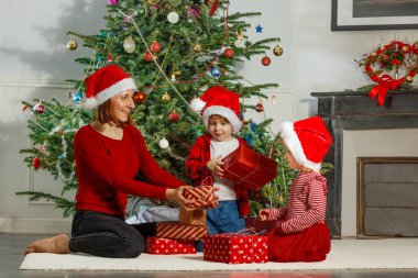 Bir kadın ve iki çocuk ile neşeli bir aile sahnesi. Şenlikli elbiseli bir kız, süslenmiş bir Noel ağacının yanında oturmuş, hediyelerini değiş tokuş ediyor.