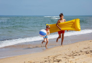 Plajda mayo giymiş iki çocuk ellerinde şişme oyuncaklarla kameraya doğru koşuyor.