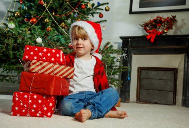 Noel Baba şapkası takan genç bir çocuk süslenmiş bir Noel ağacının önünde paketlenmiş hediyelerin yanında oturuyor.