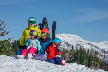 Family with two kids, mom, dad, daughter and son sit on sunny snowy slope in skiwear helmets surrounded by snow mountain ridges clipart