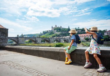Sırt çantalı ve güneş şapkalı iki çocuk, Almanya 'nın Wurzburg kentindeki yaz tatili sırasında arka planda kalan eski bir köprüyü işaret eden bir kaya çıkıntısında oturuyorlar.