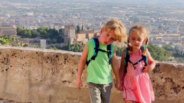 Sırt çantalı iki çocuk, Alhambra Sarayı ve Granada şehir manzarasının önünde el ele tutuşuyorlar.