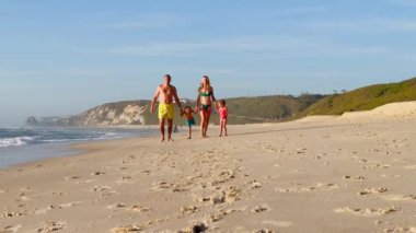 Neşeli bir aile Portekiz 'de kumlu bir plajda gün batımında arkalarında ayak izleri bırakarak neşeli bir koşu yapıyor.