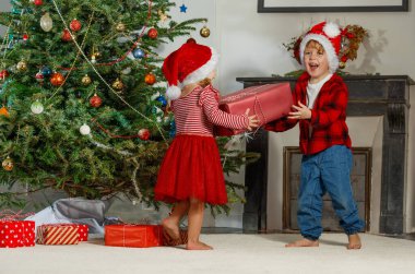 Noel Baba şapkalı iki neşeli çocuk güzel dekore edilmiş bir Noel ağacının önünde paketlenmiş bir hediye tutuyorlar.