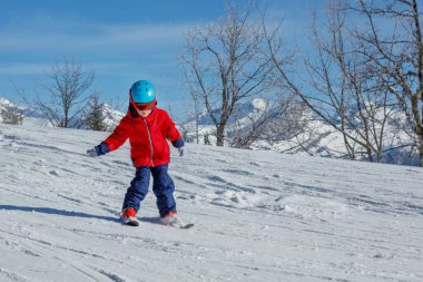 Parlak kırmızı ceketli ve koruyucu mavi kasklı genç kayakçı uzaktaki görkemli dağlarla karla kaplı bir yamaçtan aşağı iniyor.