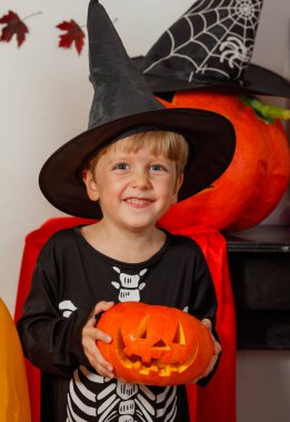 Siyah şapkalı gülümseyen çocuk elinde oyulmuş bir balkabağı, Cadılar Bayramı temalı eşyalar arasında duruyor.