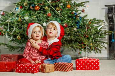 Arka planda süslü bir Noel ağacı ile, Noel Baba şapkalı iki çocuk oturur ve poz verir, hediyelerini tutar, neşeli görünürler.